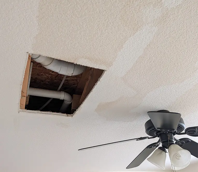 Large opening in textured drywall ceiling exposing plumbing before ceiling repair